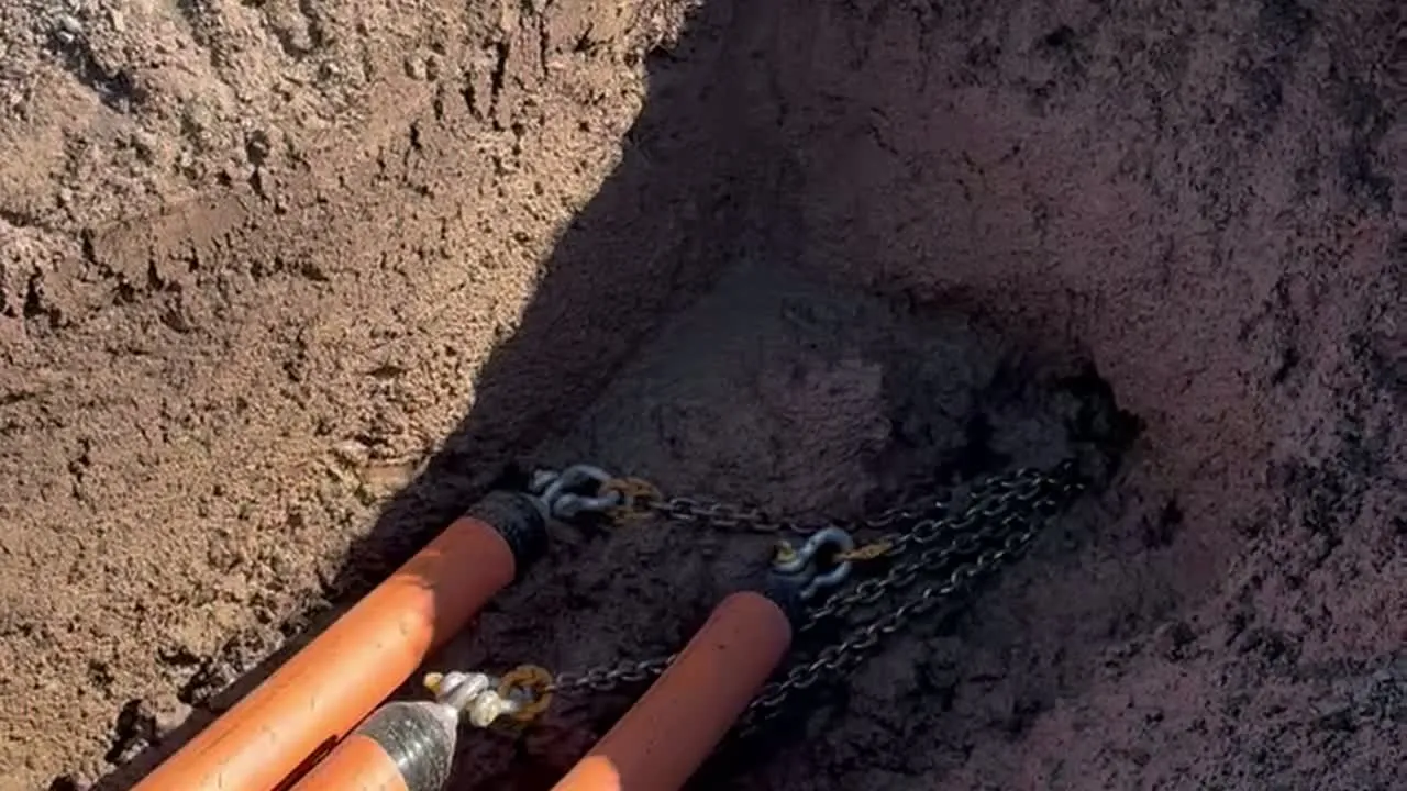 Two orange conduits with pulling grips and chains lying at the bottom of an open trench.